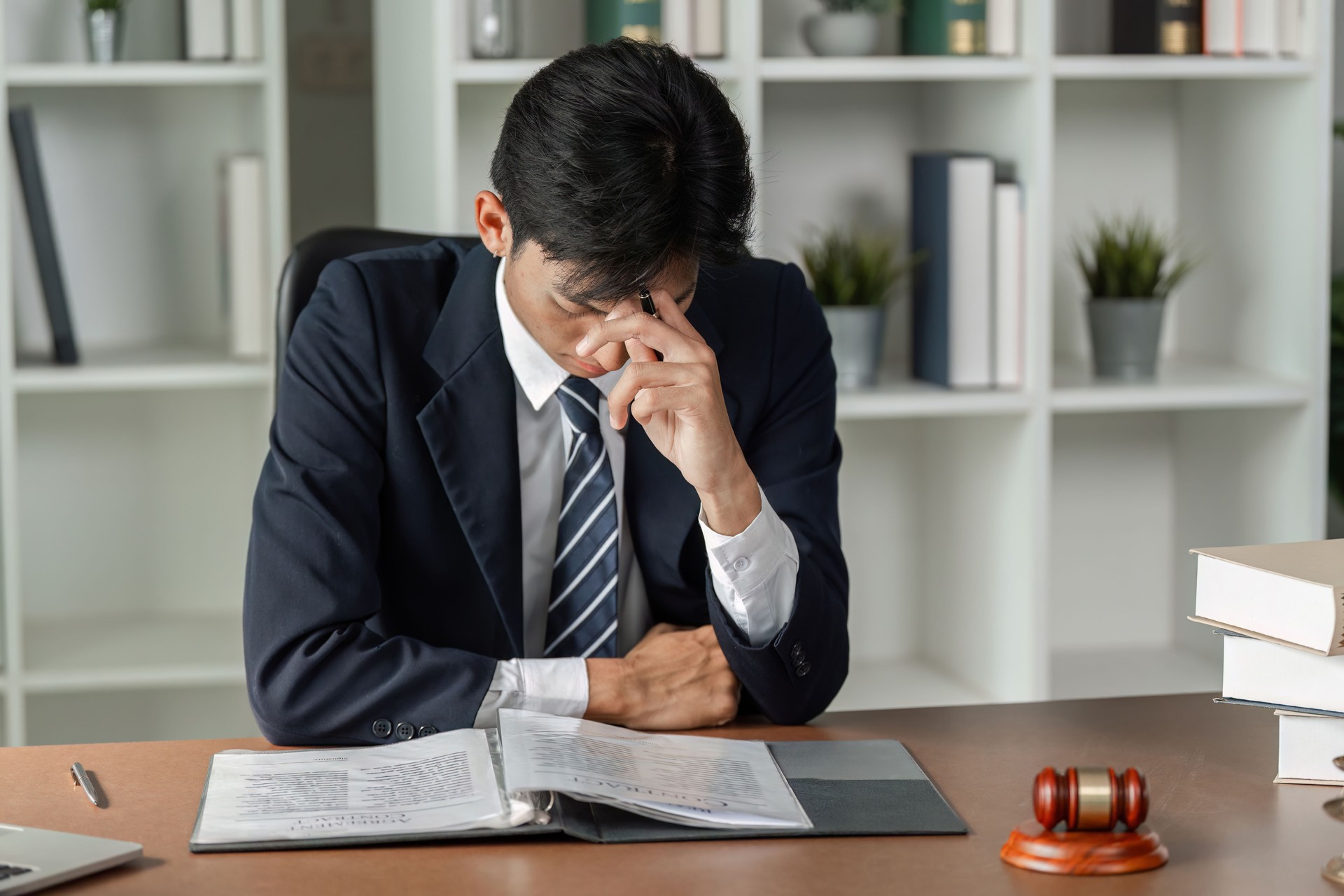 Stressed lawyer sitting at desk with head in hand, reviewing legal documents and gavel nearby.